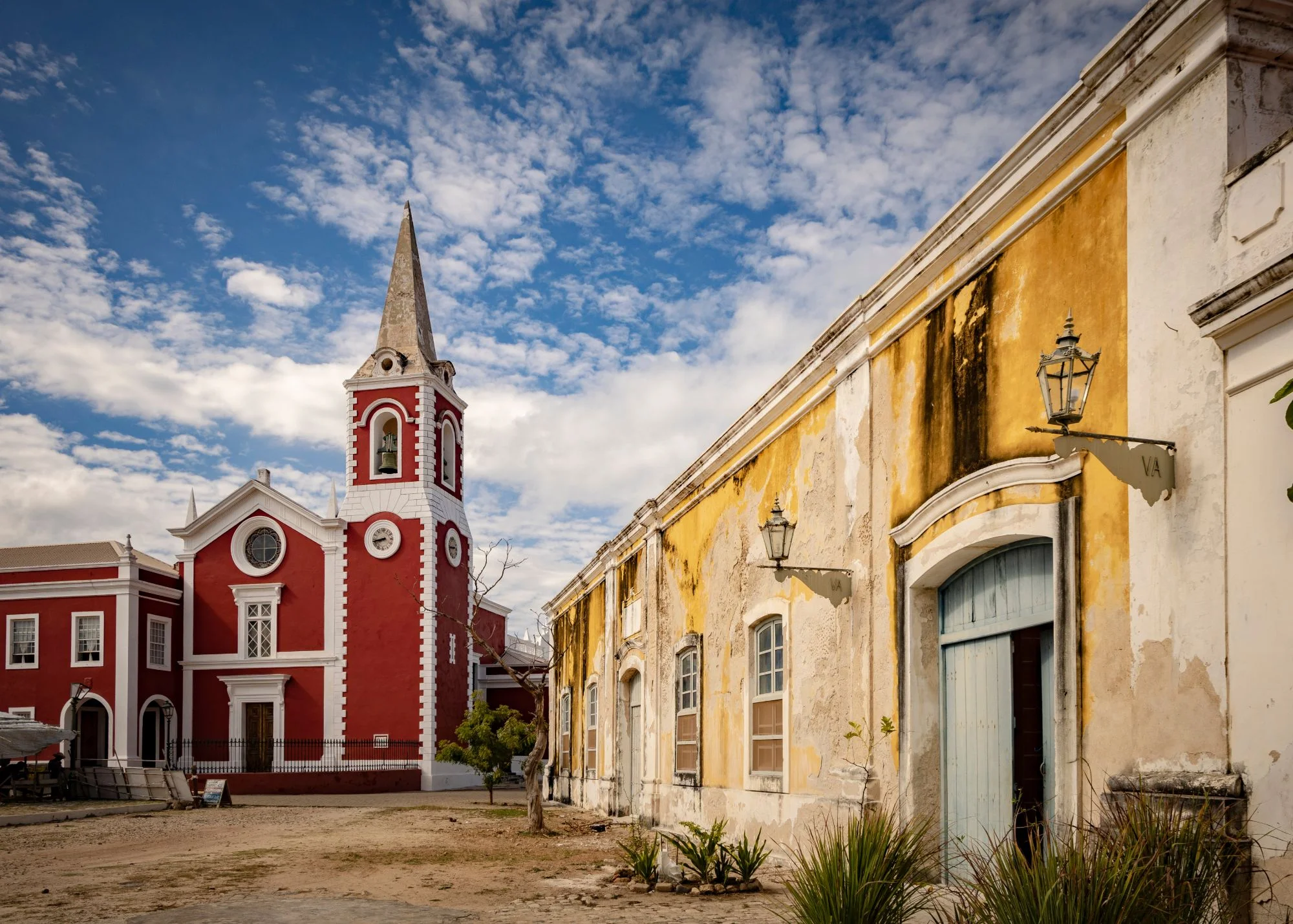 Igreja de fachada vermelha e branca com campanário alto, ao lado de um edifício amarelo e branco com portas azuis, sob um céu azul com nuvens