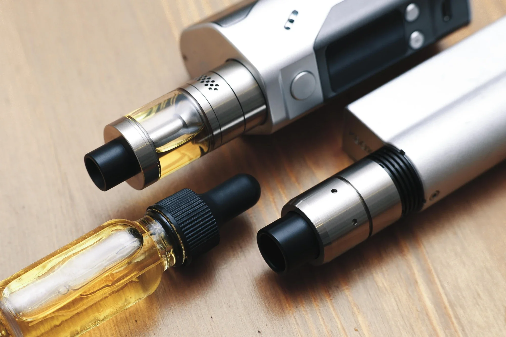 Dois cigarros eletrónicos de metal prateado e um frasco de líquido amarelo com conta-gotas, pousados sobre uma mesa de madeira
