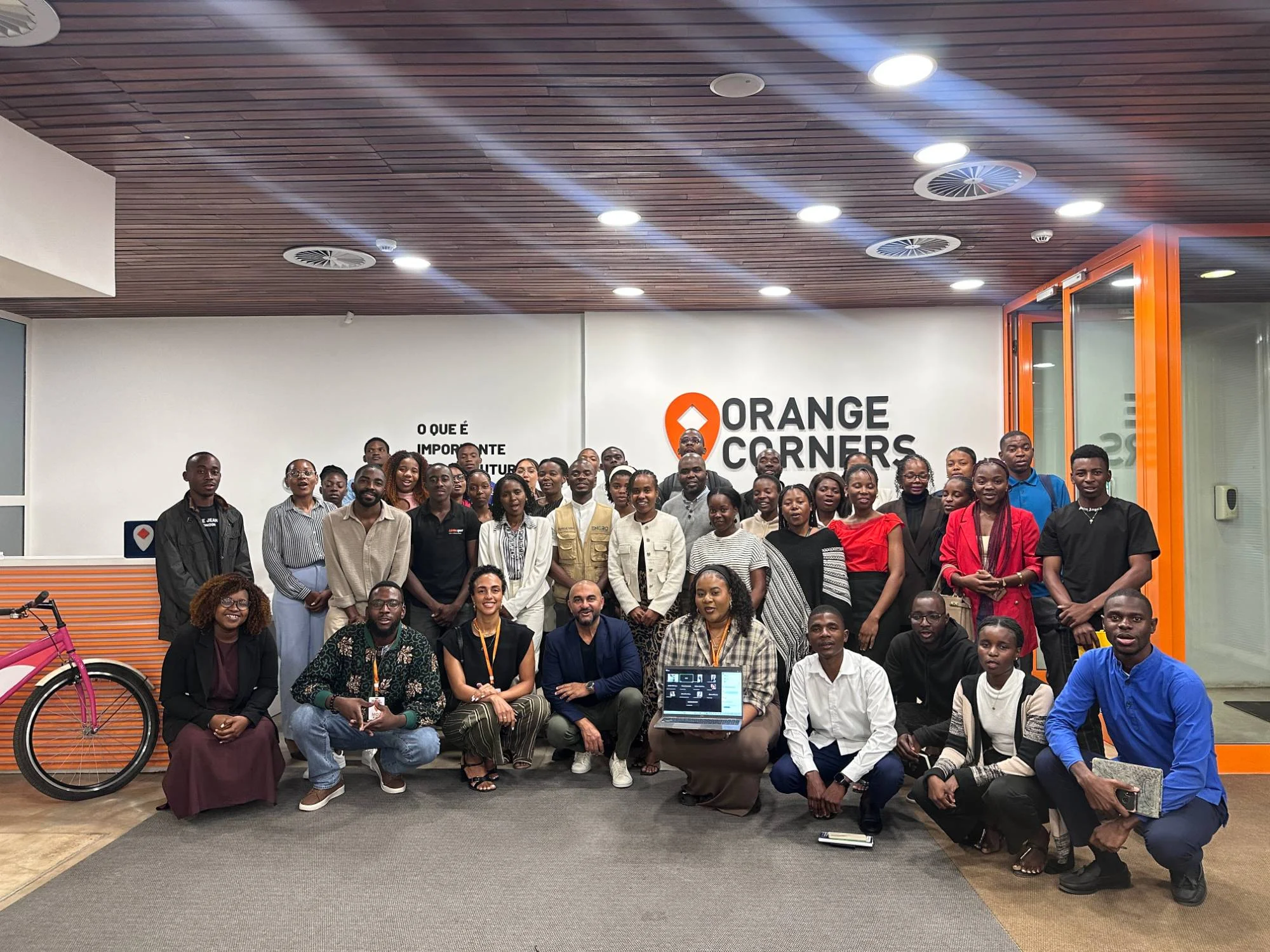 Grupo de pessoas de pé e agachadas num espaço com paredes brancas, tecto de madeira e o logótipo Orange Corners ao fundo, junto a uma bicicleta cor-de-rosa